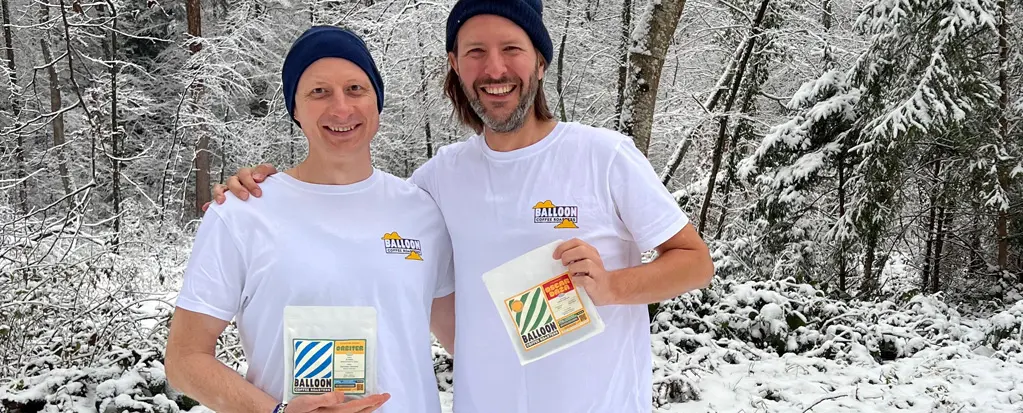 Zwei Männer mit weißen T-Shirts und Wintermützen stehen im verschneiten Wald und halten Produkte in den Händen